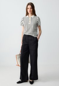 Femme portant un pantalon large noir, un polo à manches courtes noir et blanc à rayures, tenant un tote bag beige avec des bords marron, se tenant contre un fond blanc.