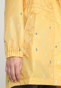 Gul vattentät jacka med regndroppsmönster i blått och vitt. Har elastiska muddar, sidoficka och gula tryckknappar.