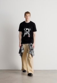 T-shirt preto com um design gráfico de um cão na frente, combinado com calças largas bege e uma camisa cinza com padrão xadrez atada à cintura.