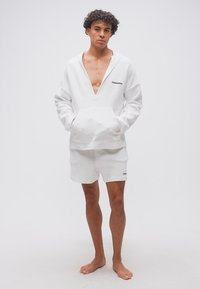 Νεαρός άνδρας, ξυπόλητος, φοράει λευκή υφασμάτινη φούτερ με βαθύ V-λαιμόκοψη και ταιριαστό σορτς, στέκεται με τα χέρια στις τσέπες σε απλό φόντο.