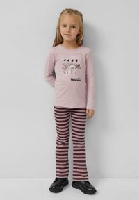 Hellrosa Langarmhemd mit grafischen Tänzern und dem Text "Du kannst mich Prinzessin nennen". Gestreifte kastanienbraune und rosa Schlaghosen. Schwarze Schuhe.