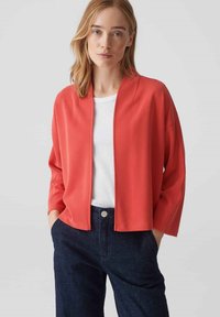 Veste rouge à coupe décontractée avec un devant ouvert, un design décontracté, une texture lisse et des manches trois-quarts, portée sur un haut blanc et un jean foncé.