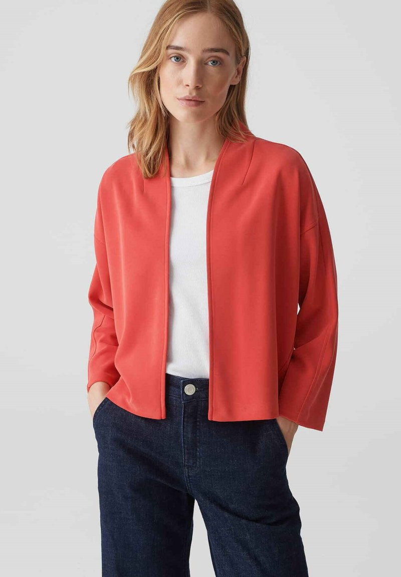 Veste rouge à coupe décontractée avec un devant ouvert, un design décontracté, une texture lisse et des manches trois-quarts, portée sur un haut blanc et un jean foncé.
