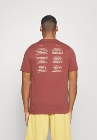 Bordo puuvillatee, mille tagaküljel on valge trükk linnade nimedest, korraldatud ida- ja lääneregioonide kaupa. Klassikaline ümar kaelus.