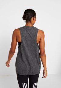 Šedý bezrukávový sportovní top s volným střihem, kulatým výstřihem a širokými rameny, kombinovaný s černými legínami se bílými pruhy.