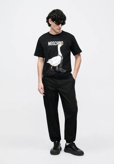Čierne tričko Moschino s bielou grafikou kačky s bodkovanou kravatou, kombinované s čiernymi nohavicami a čiernymi teniskami.