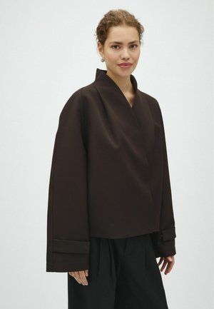 Femme aux cheveux bouclés portant une veste ample marron foncé et un pantalon noir, debout devant un fond clair uni.