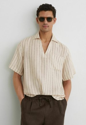 Mann mit brauner Sonnenbrille, cremefarbenem kurzärmeligem Hemd mit vertikalen Streifen und dunkler Tunnelzughose, stehend vor einer schlichten weißen Wand.