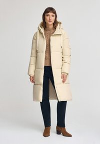 Kurtka puchowa w kolorze beżowym, sięgająca do kolan, z kapturem i sznurkami, z przodu zapinana na guziki, pikowana. Noszona z brązowym swetrem z dzianiny i ciemnymi spodniami.