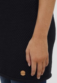 Czarny materiał o fakturze z tkanym wzorem, z okrągłym drewnianym akcentem z logo przy krawędzi oraz zbliżeniem na dłoń opartą o tkaninę.