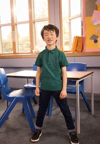 Grøn polo med korte ærmer og et rødt broderet logo, kombineret med mørkeblå jeans og sorte sneakers, i et klasseværelse.