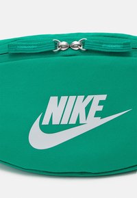 Grön tygmidjeväska med en framträdande vit Nike-logotyp, dragkedjestängning och strukturerat material med en slät yta.