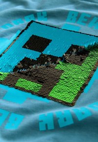 Grafisk t-shirt med et pixeliseret design i blå, sort og grønne palietter på en lys blå stoffet med iøjnefaldende tekstakcenter.