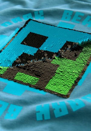 Grafická trička s pixelovaným designem v modré, černé a zelené flitry na světle modré látce s výrazným textovým akcentem.