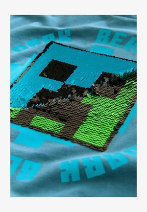 Graafiline t-särk, millel on pikseliline disain siniste, mustade ja roheliste sädelevate detailidega helesinises kangas ning julged sõnumid.