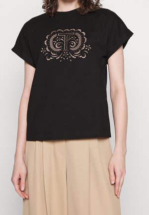 Person mit schwarzem Kurzarmhemd mit detailliertem Spitzendesign in Blumenform auf der Brust und beigen, hoch-taillierten, plissierten Hosen.