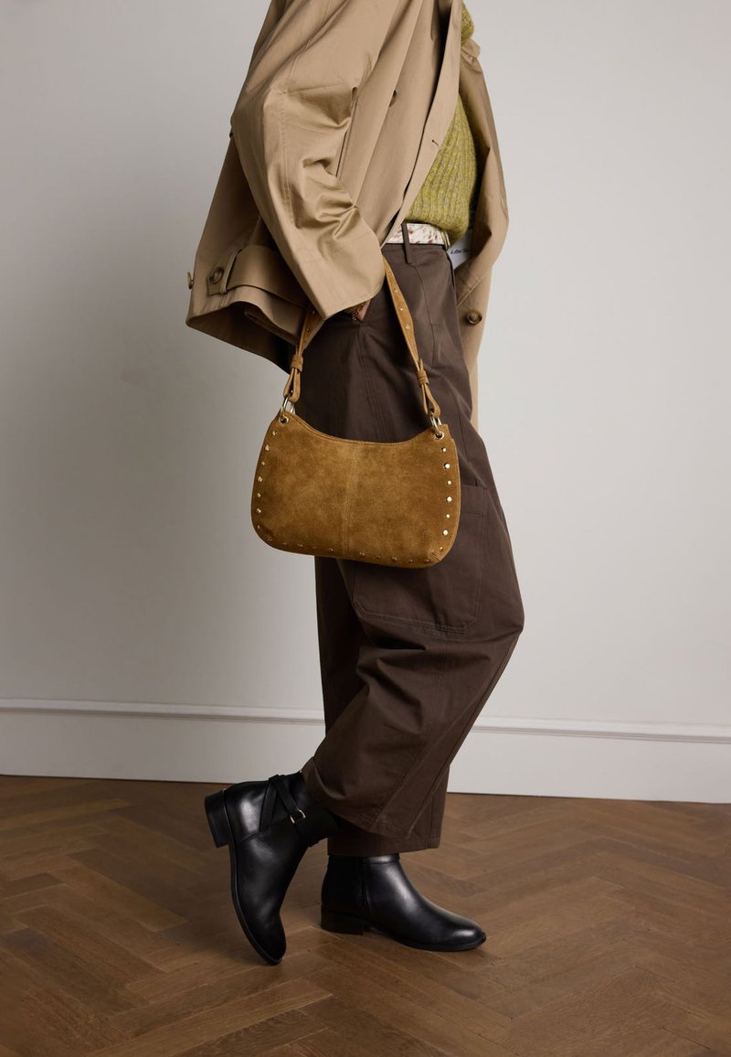 Borsa a tracolla in suede marrone con dettagli borchiati, forma curvata. Indossata con un cappotto beige e stivaletti neri, su un pavimento di legno.