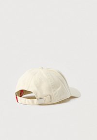 Béžová bavlnená šiltovka s oblým šiltom, nastaviteľným remienkom, strieborným kovaním a červenou značkou viditeľnou na zadnej strane.