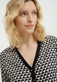 Bluzka w wzory z czarno-białym deseniem, z abstrakcyjnymi kształtami. Dekolt w kształcie litery V oraz akcenty w postaci guzików podkreślają zorganizowany wygląd.