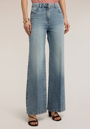 Donna che indossa jeans a gamba larga a vita alta di colore azzurro chiaro, sandali con tacco marroni con cinturini e braccialetti dorati al polso sinistro.