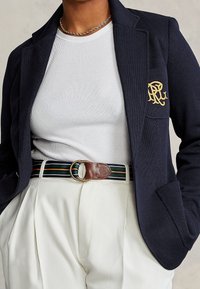 Personne portant une chemise blanche côtelée, un blazer bleu marine avec une broderie dorée sur la poche, un pantalon blanc taille haute, une ceinture rayée et un collier chaîne.