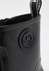 Botte cheville en caoutchouc noir avec une finition lisse, dotée d'un détail logo circulaire, de panneaux latéraux élastiques et d'une languette pour un enfilage facile.