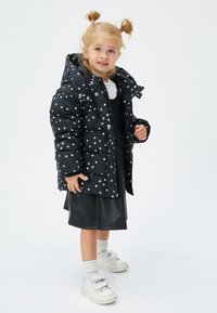 Sort puffjakke med hvide prikker, lynlås foran, stor hætte. Båret over en sort kjole, sammen med hvide sokker og sneakers.