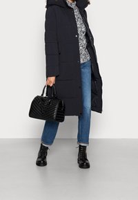 Abrigo acolchado negro con capucha, top gris estampado, vaqueros azules, bolso negro acolchado y botas negras con cordones. Texturas suaves y diseño en capas.