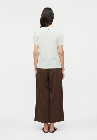 Άτομο με μακριά σκούρα μαλλιά, που φοράει ανοιχτόχρωμο κοντομάνικο πουκάμισο, φαρδιά καφέ παντελόνια και μαύρα σανδάλια, στέκεται γυρισμένο με την πλάτη σε λευκό φόντο.