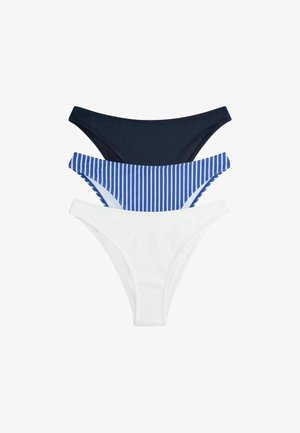 Donna in piedi scalza contro un muro bianco, che indossa un costume da bagno bianco a due pezzi con top con scollo quadrato e slip a vita alta.