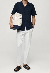 Marineblaues kurzärmeliges Hemd, weiße schmal geschnittene Hose, schwarze Sandalen und eine cremefarbene Tasche mit marineblauen Akzenten. Glatte Stofftexturen, lässiges Design.