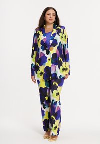 Femme debout portant un blazer et un pantalon assortis à motif floral en bleu, violet, jaune et blanc, avec des talons ouverts jaunes, sur fond blanc.