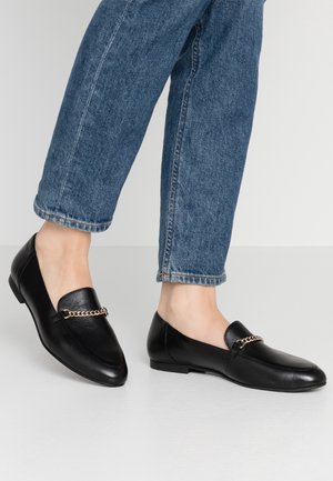 Mocassins en cuir noir avec détail chaîne dorée portés avec un jean bleu retroussé sur un sol blanc et un fond gris clair.