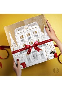 Boîte cadeau ornée d'une illustration détaillée d'un immeuble parisien en or et blanc, avec un ruban rouge et un message. Texture lisse, forme rectangulaire.