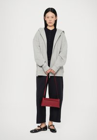 Grijze oversized hoodie met een voorrits, marineblauw overhemd met knopen, zwarte wijde broek en zwarte platte schoenen. Een kleine rode crossbody tas is toegevoegd.
