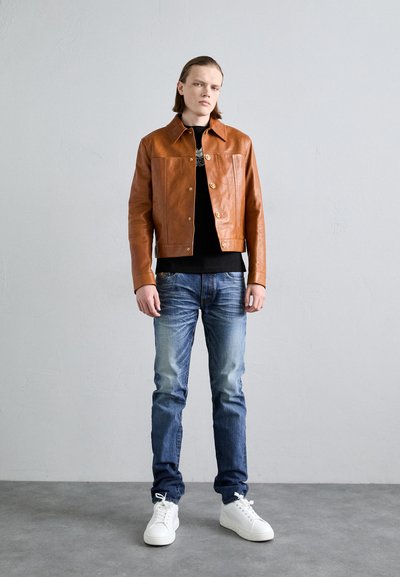 Braune Lederjacke mit Knopfverschluss, schwarzes Hemd, blaue Jeans und weiße Sneaker. Die Jacke hat ein strukturiertes Design und einen Kragen.