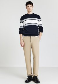 Junger Mann, der einen navyblauen, weißen und grauen gestreiften Pullover, beige Hosen und schwarze Schnürschuhe trägt und mit einer Hand in der Tasche steht.