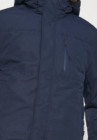 Chaqueta aislante de color azul marino con cuello alto, cremallera frontal y bolsillo en el pecho. Presenta una textura de tela suave y capucha ajustable.