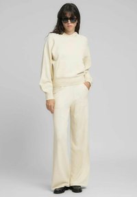 Sudadera con capucha de color crema con bolsillo frontal, combinada con pantalones de pierna ancha a juego. El conjunto presenta una textura suave y puños acanalados. Las gafas de sol negras añaden contraste.