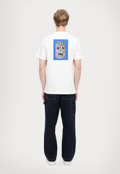 Hvit bomulls t-skjorte med et blått rektangulært grafikk som har et teksturert svart design på baksiden, kombinert med mørke denimbukser.