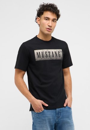 Giovane uomo con capelli ricci che indossa una t-shirt nera con il logo Mustang e jeans blu, in piedi con le mani in tasca davanti a uno sfondo semplice.