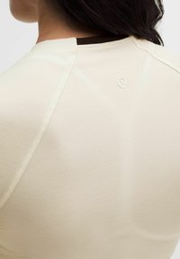 Camiseta deportiva de color beige claro con una textura suave, mangas raglán y un pequeño logo en la parte posterior. Cuenta con un ribete de color marrón en el cuello a contraste.