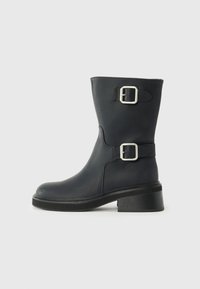 Botas de cuero negras hasta el tobillo con dos hebillas plateadas, puntera redondeada y tacón grueso. La textura es lisa con un ligero brillo.