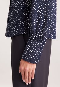 Mano accanto a pantaloni scuri, indossando una blusa blu navy con pois bianchi e polsini arricciati con bottone.