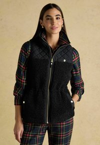 Czarna, pikowana kamizelka z puszystym, teksturowanym przodem, z bocznymi kieszeniami i złotym zamkiem błyskawicznym. Noszona na koszulę w kratę i dopasowane spodnie.