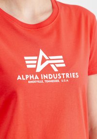 Raudona medvilninė marškinėliai su balta "Alpha Industries" logotipu ir sparnais, o apačioje tekstas su vieta. Trumpomis rankovėmis ir apvalia iškirpte.