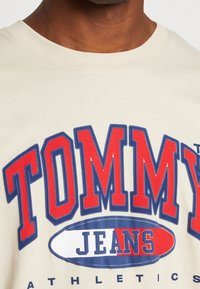 Beige bavlněné tričko s kulatým výstřihem, které má velké červené a modré logo "TOMMY JEANS" s bílým oválným akcentem a modrými švy.