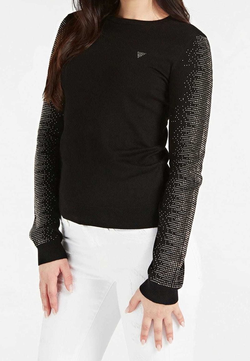 Femme portant un haut noir à manches longues avec des motifs de points argentés sur les manches et un pantalon blanc sur un fond blanc uni.