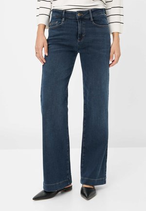 Kvinde iført mørkeblå bredbenede jeans, sorte flade sko med spids tå og en hvid langærmet top med sorte striber.