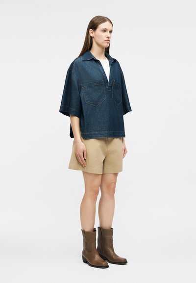 Γυναίκα που στέκεται φορώντας μια φαρδιά σκούρα τζιν κοντομάνικη μπλούζα, μπεζ σορτς και καφέ μποτάκια μέχρι τα μέσα της γάμπας σε απλό λευκό φόντο.